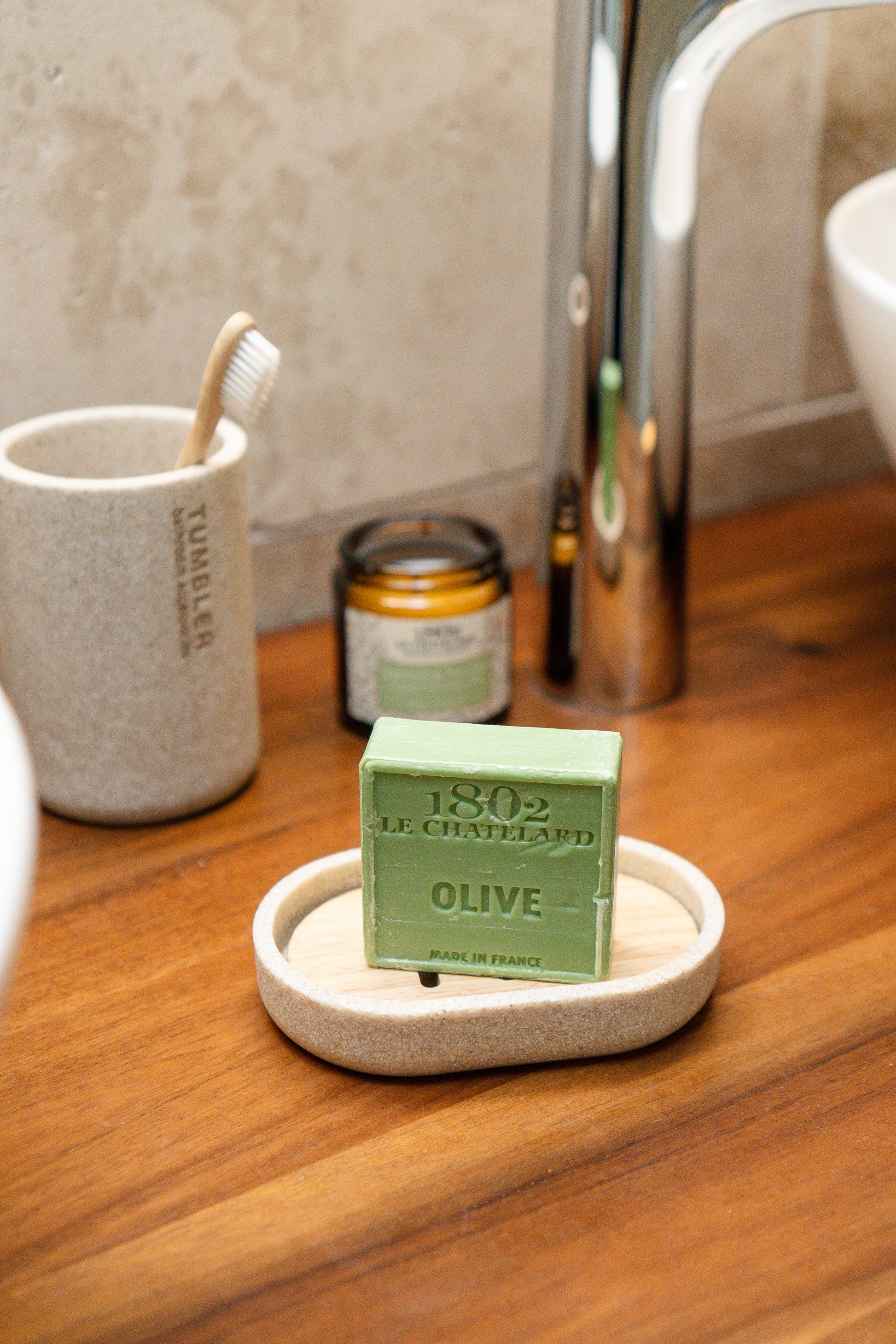 Green bar of soap on a wooden bathroom counter with a toothbrush holder and candle in the background.