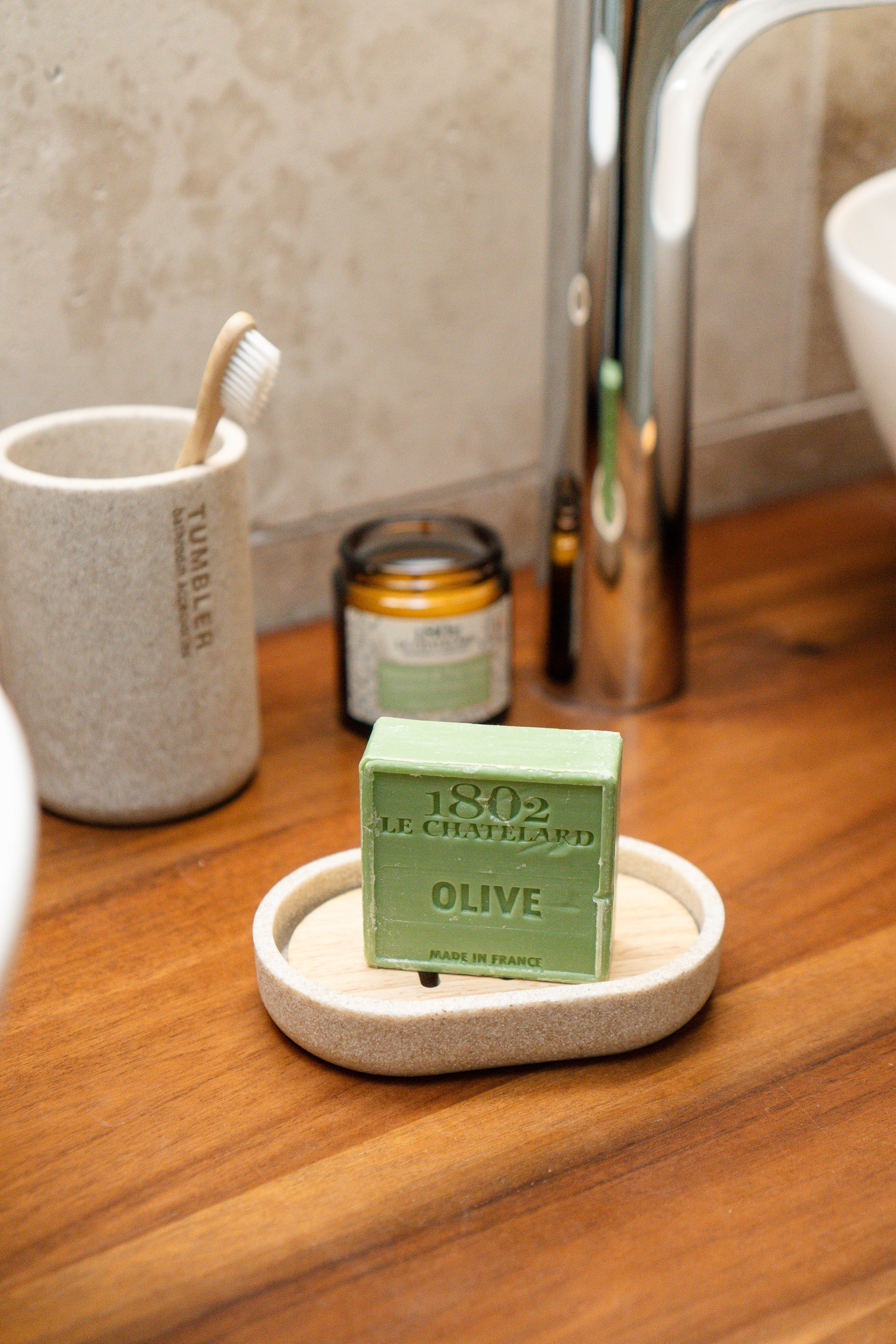 Green bar of soap on a wooden bathroom counter with a toothbrush holder and candle in the background.