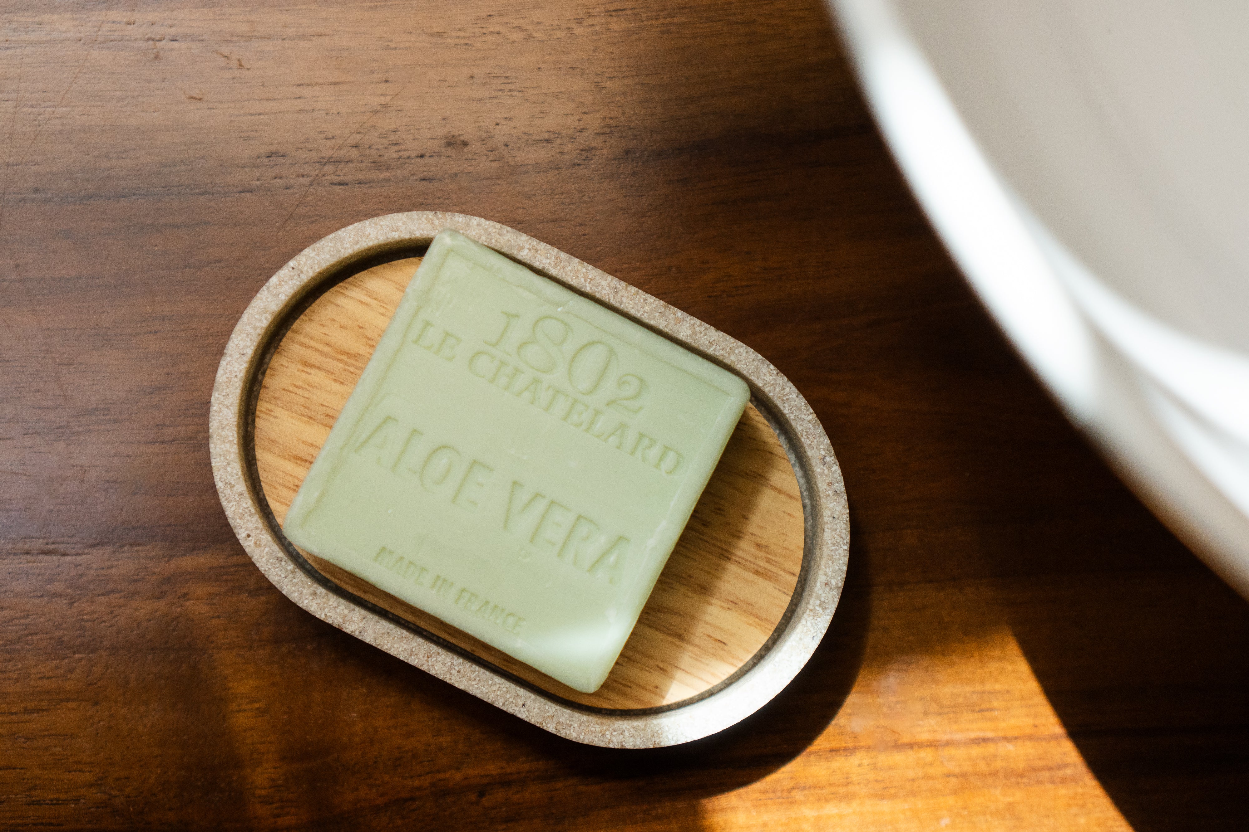 Green bar of soap on a wooden soap dish with '1802 Le Gentilhomme' branding on a wooden surface.